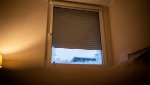Fenêtre d’un appartement en hiver, volet roulant manuel partiellement fermé, lumière intérieure chaleureuse et légère buée sur la vitre.